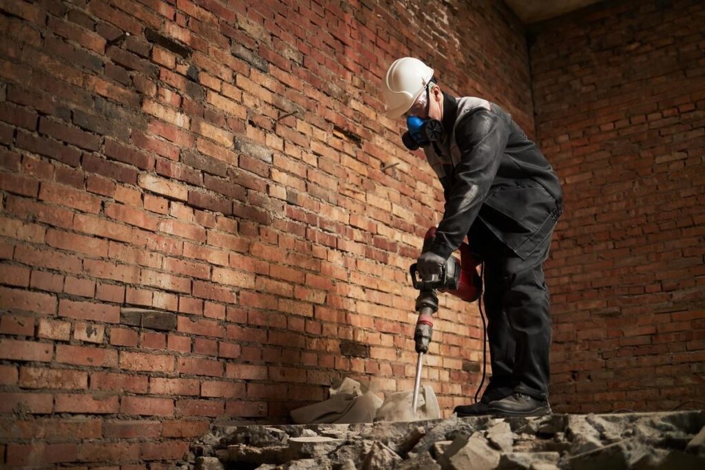 A person wearing protective gear uses a jackhammer to break concrete rubble inside a building with exposed brick walls.