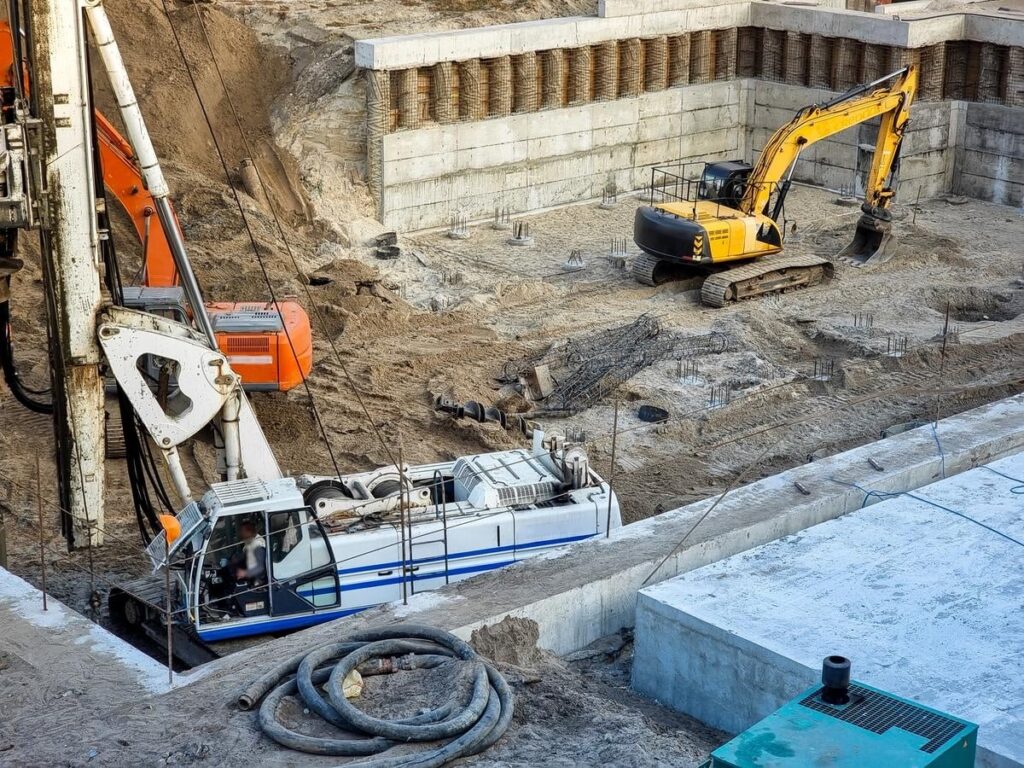 Excavators and construction machinery operate at a building foundation site with exposed concrete walls and rebar.