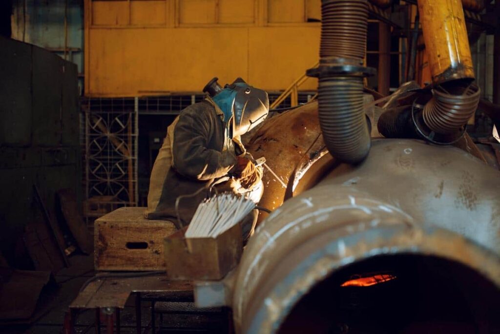 A person wearing protective gear welds metal parts together in an industrial workshop with machinery and tools visible.