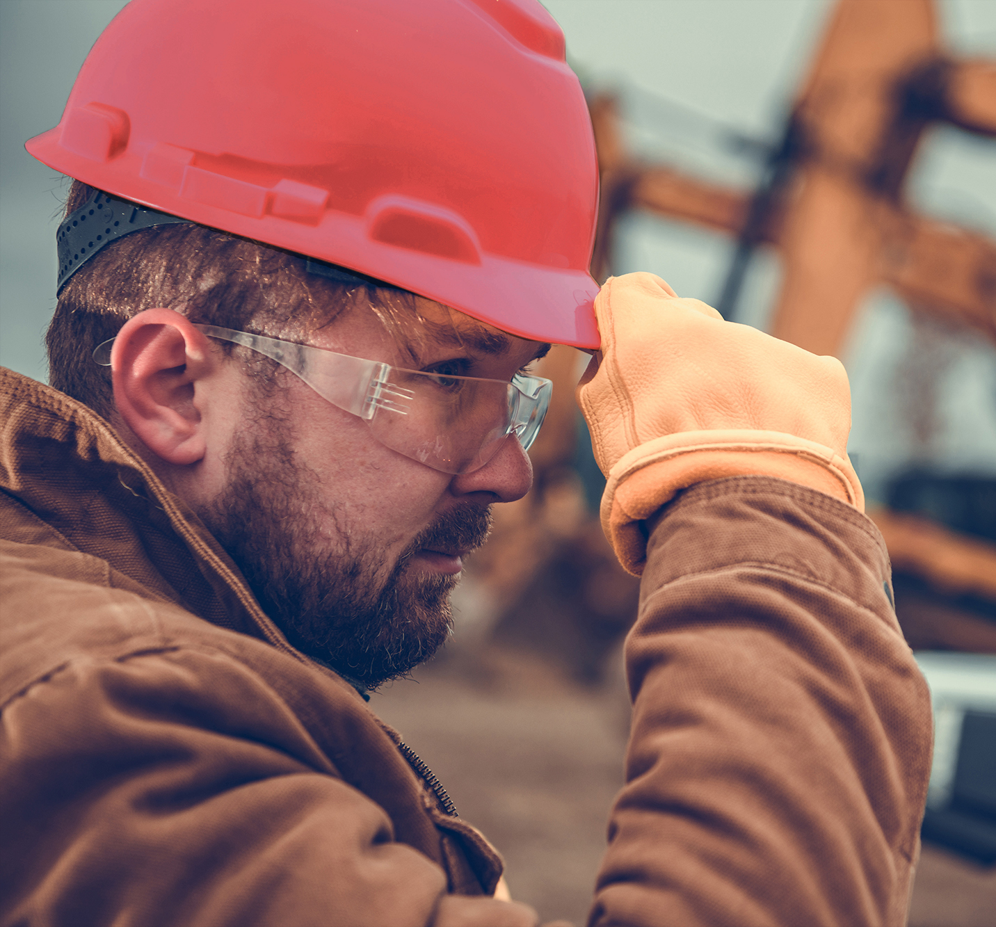 A man wearing a red hard hat, safety glasses, brown jacket, and orange gloves is adjusting his helmet at a construction site.