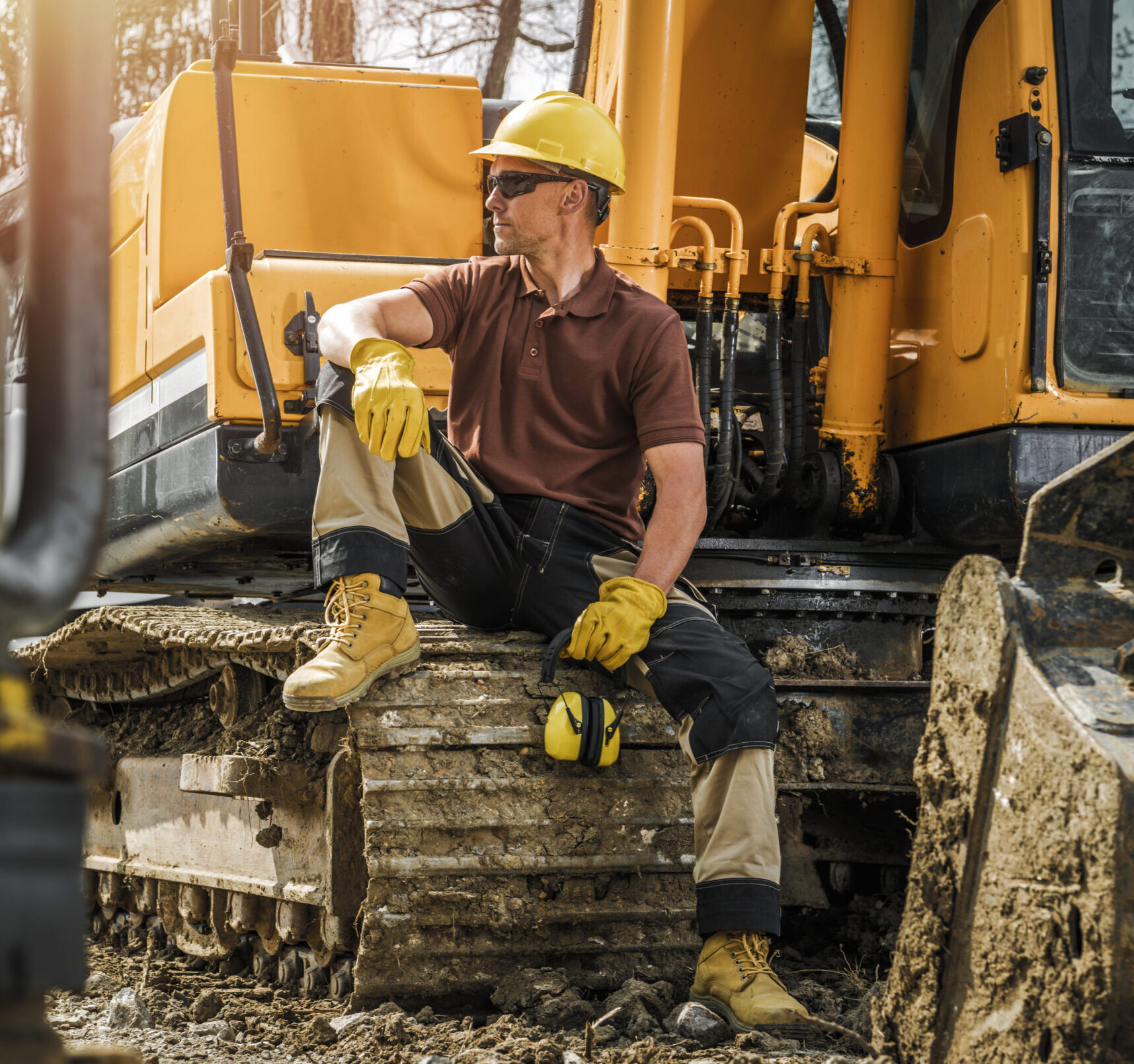 Construction worker in safety gear sits on the track of a muddy excavator, holding ear protection and looking to the side at a construction site.